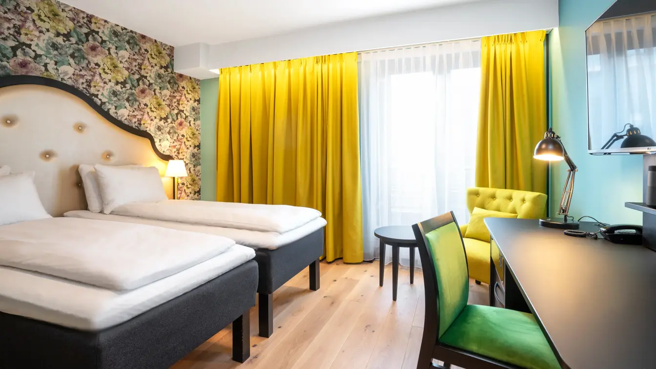 To enkeltsenger, blomstret tapet bak sengene, gul stol, arbeidspult og store vinduer med store gule gardiner på twin room på Thon Hotel Cecil i Oslo sentrum