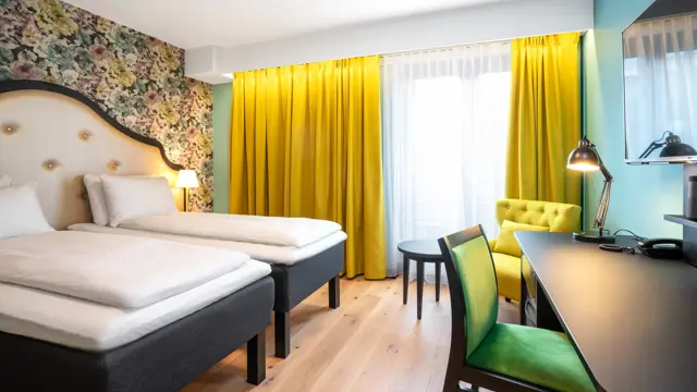 To enkeltsenger, blomstret tapet bak sengene, gul stol, arbeidspult og store vinduer med store gule gardiner på twin room på Thon Hotel Cecil i Oslo sentrum