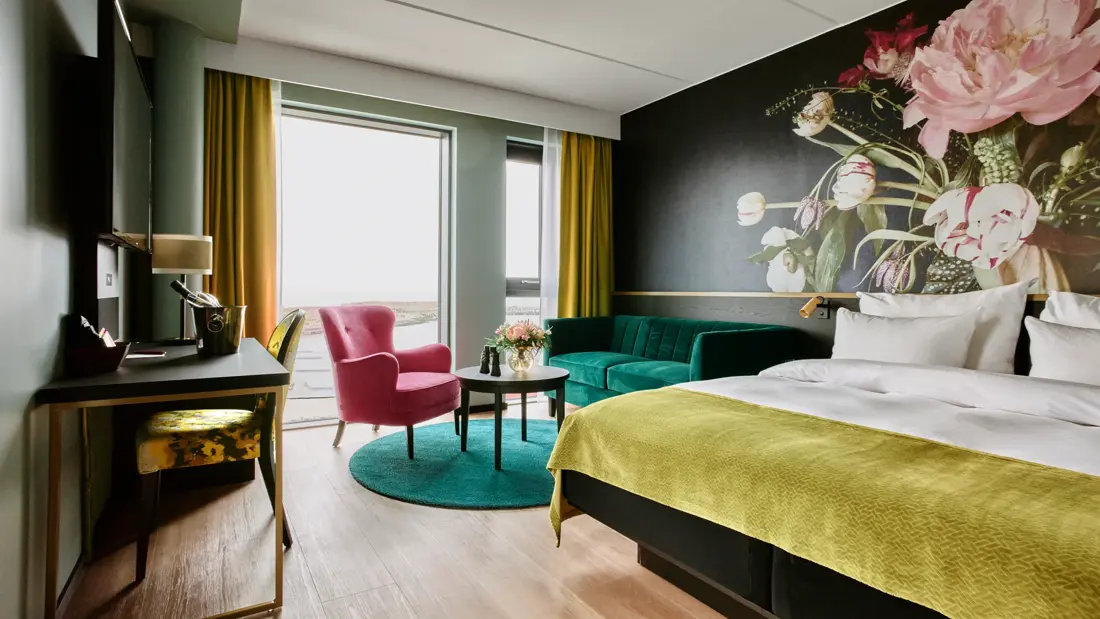 Blomstertapetvegg, dobbeltseng, grønn sofa, Vindu, gule gardiner, skrivebord, grønne vegger, rosa lenestol, TV