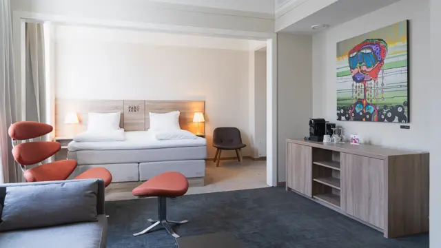 Suite med dobbeltseng, rød lenestol med puff, kommode, teppe på gulvet, fargerikt bilde på veggen