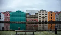 Utsnitt av Kjøpmannsgata som speiler seg i Nidelva, med nr. 15 i midten. I forgrunnen en benk.