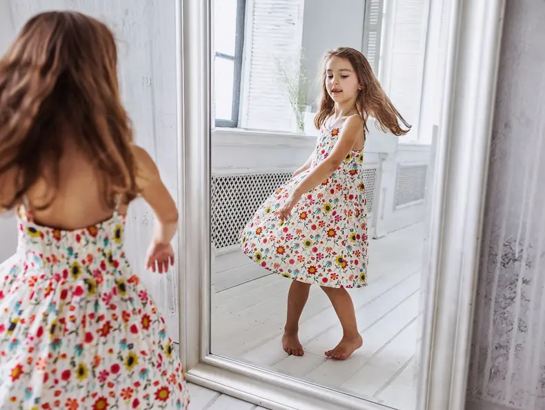 lovely smiling little girl about 6-10 years old on the floor in cute flower summer dress smiling  turning around at the mirror