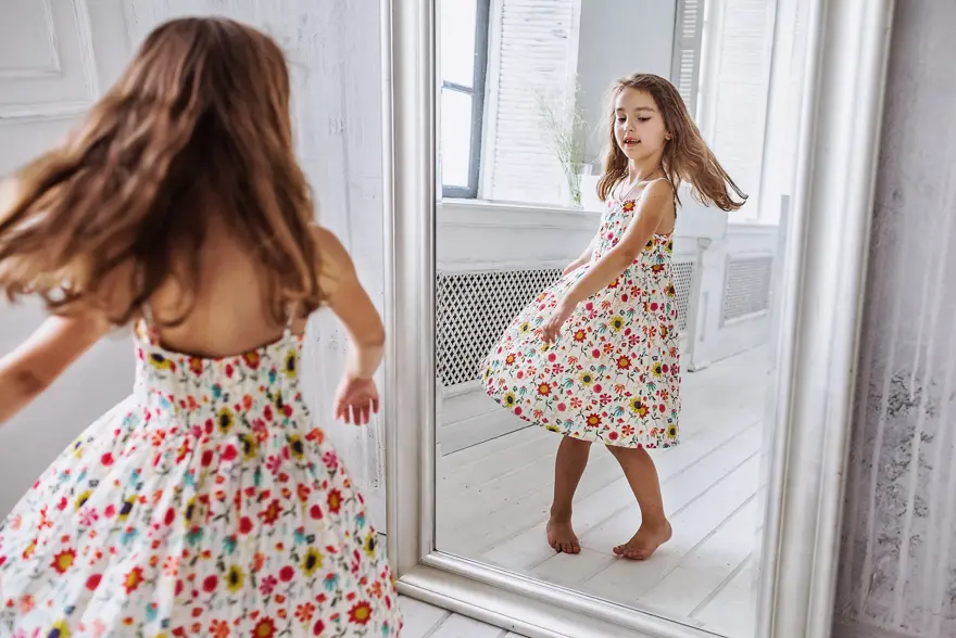 lovely smiling little girl about 6-10 years old on the floor in cute flower summer dress smiling  turning around at the mirror
