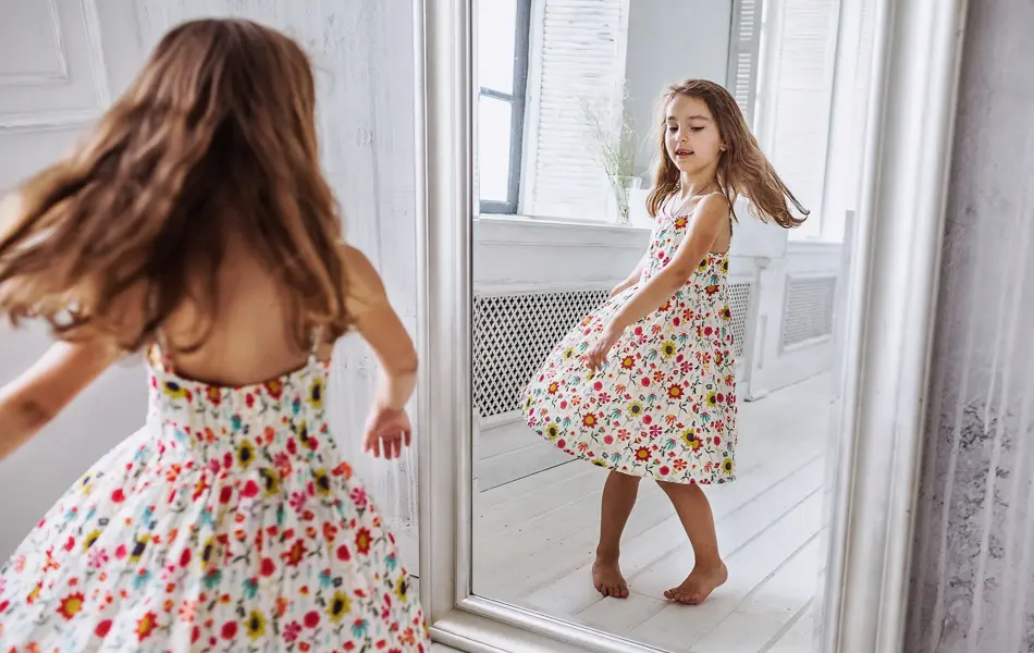 lovely smiling little girl about 6-10 years old on the floor in cute flower summer dress smiling  turning around at the mirror