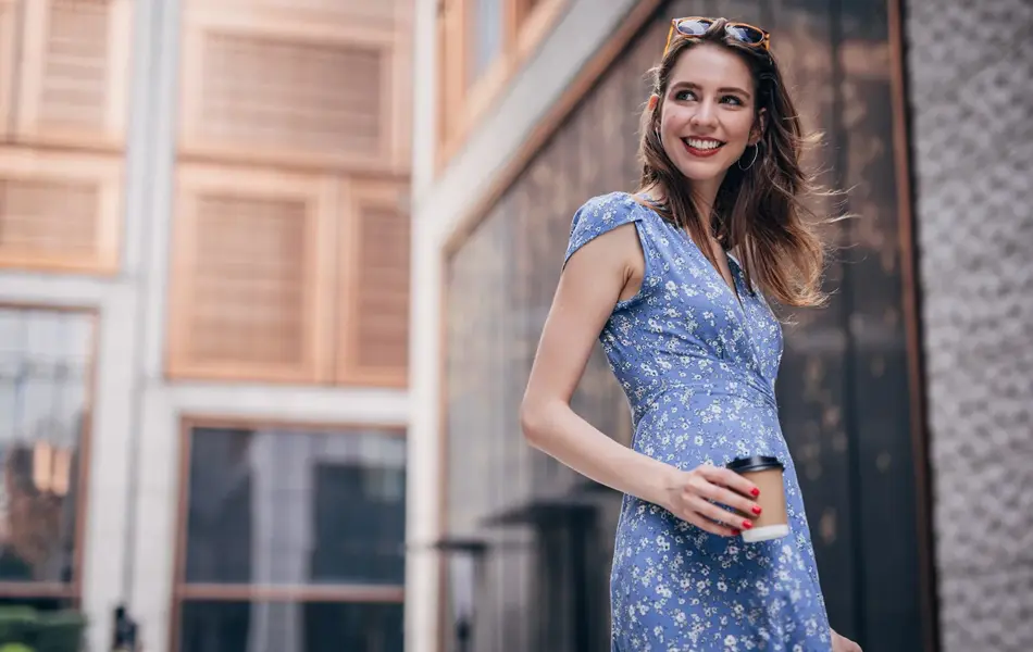 Dame iført blå blomstrete kjole, solbriller på hodet og rød neglelakk med en take away-kaffe i hånden. 