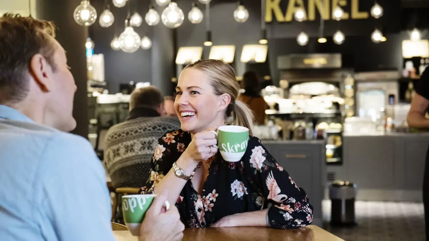 Venke Knutson sitter ved et bord og drikker kaffe fra en grønn koppe med teksten Skravlekopp på.