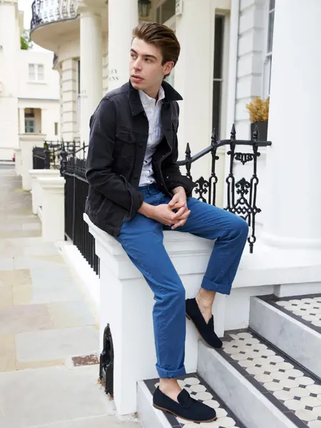 Mann med blå jakke, blå bukse og mørkeblå loafers sitter på gelender ved trapp utendørs 