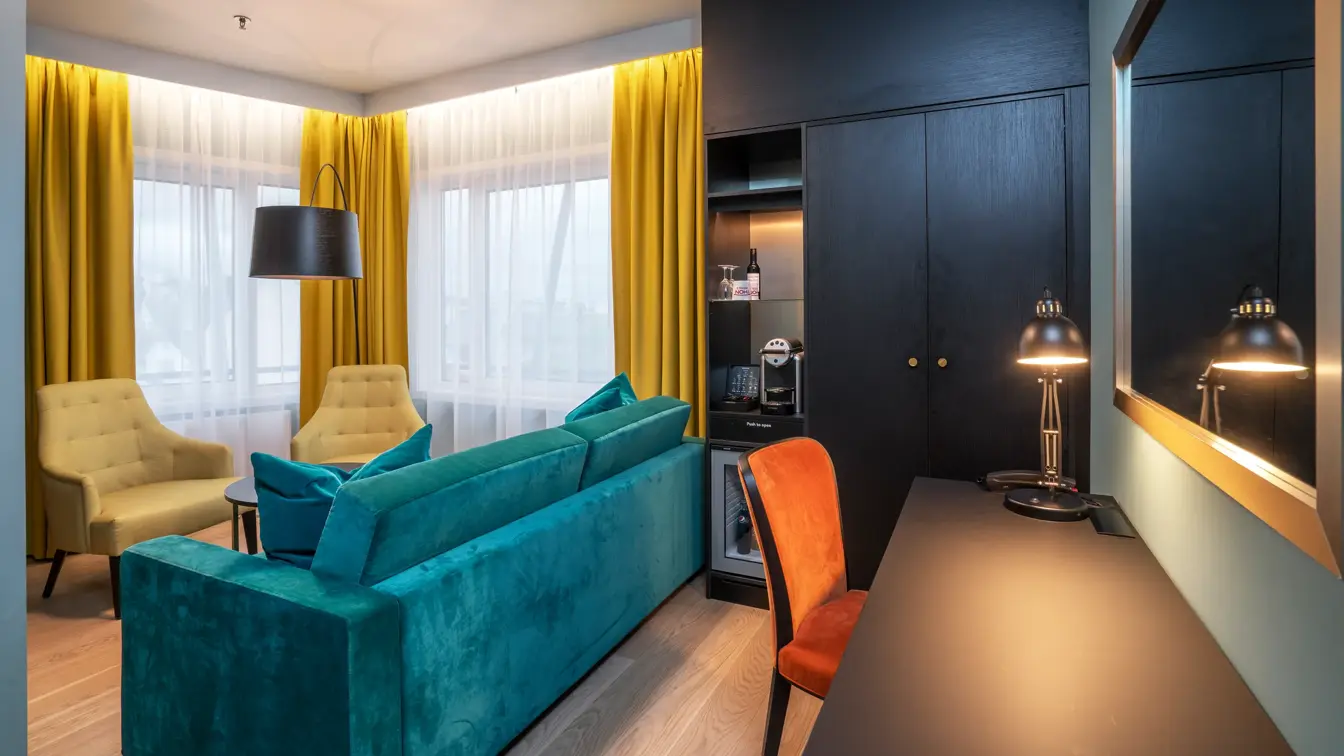 To store vinduer med gule gardiner, to gule stoler, en grønn toseter sofa, stort svart veggskap, svart skrivebordspult med en oransje stol. 