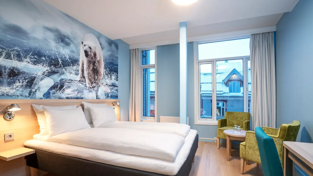 Tapetbilde med isbjørn på veggen over sengen, turkise vegger, store vinduer, dobbeltseng, sittegruppe med to stoler og bord, skrivebord med stol i dobbeltrom på Thon Hotel Polar i Tromsø.