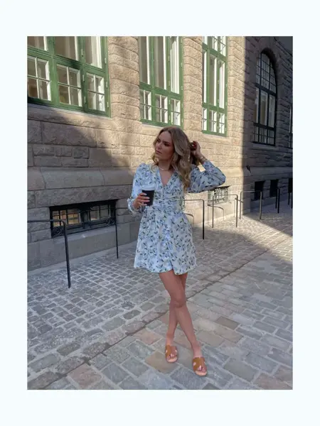 Ung dame står ute i en by med kort blomstrete kjole og kaffe. 