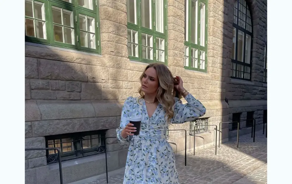 Ung dame står ute i en by med kort blomstrete kjole og kaffe. 