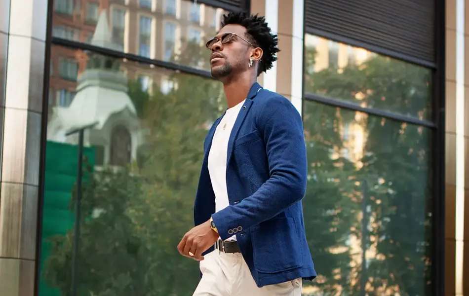 En mann i full lengde fra siden går utenfor og har på seg trendy forretningsklær, blå jakke, hvite bukser og loafersko. Stilig mannlig motemodell, Street style