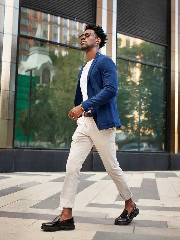 En mann i full lengde fra siden går utenfor og har på seg trendy forretningsklær, blå jakke, hvite bukser og loafersko. Stilig mannlig motemodell, Street style