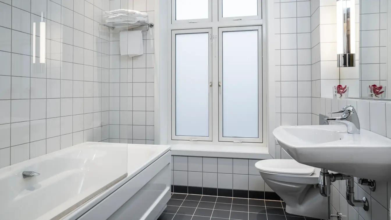 Vegger med hvite fliser, vindu med frostet foliering, hvitt badekar med glassvegg, hvit servant og speil. I baderom på business rom Thon Hotel Høyers i Skien.