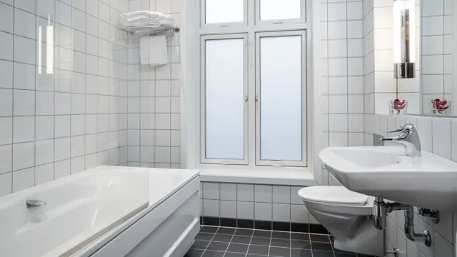 Vegger med hvite fliser, vindu med frostet foliering, hvitt badekar med glassvegg, hvit servant og speil. I baderom på business rom Thon Hotel Høyers i Skien.