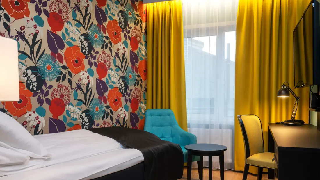 Enkel seng, blomstrende veggtapet, gule gardiner, blå og gul stol og svart pult.