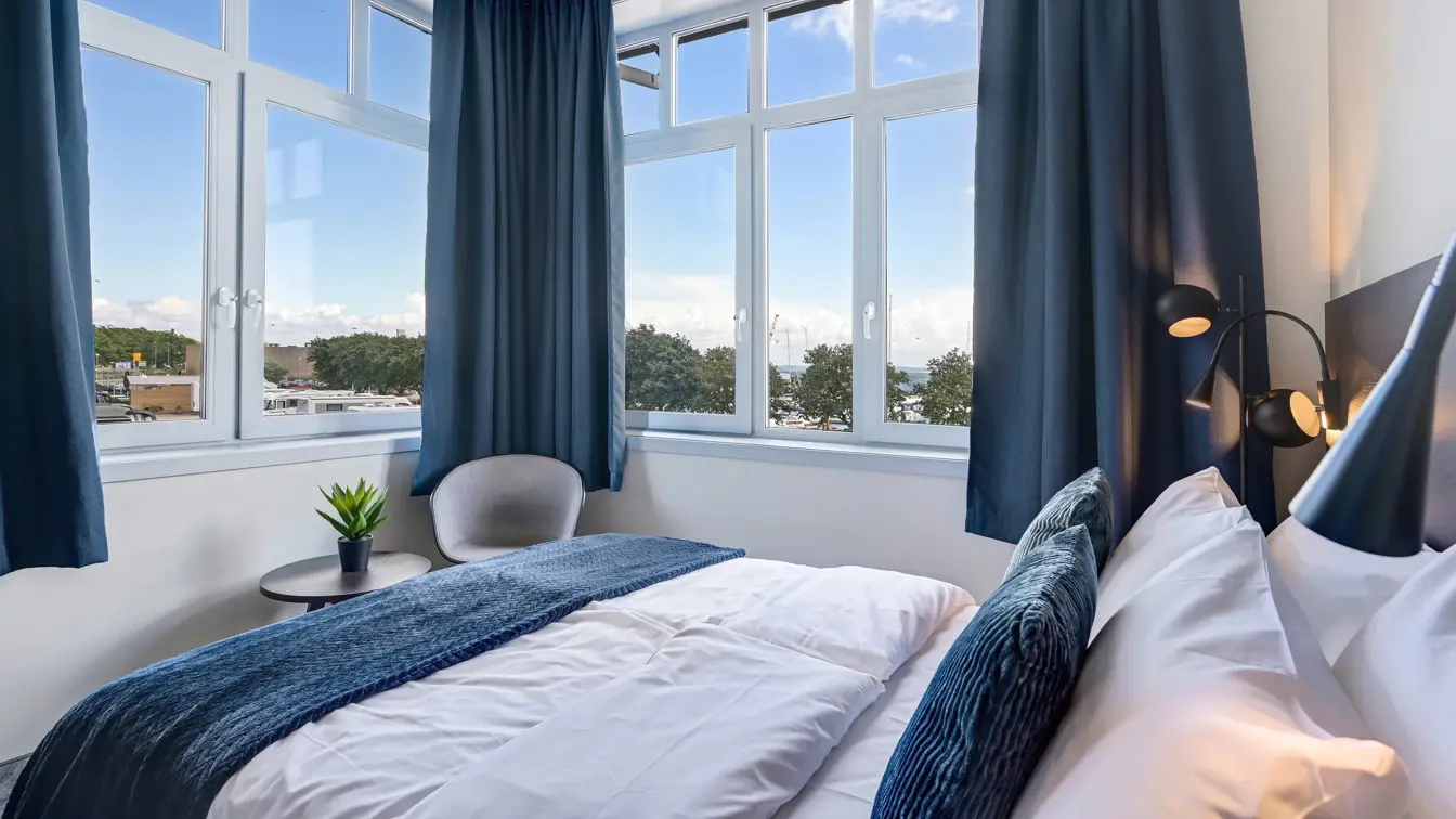 Dobbeltseng, hvite vegger, vindu, blå gardiner, stol, bord