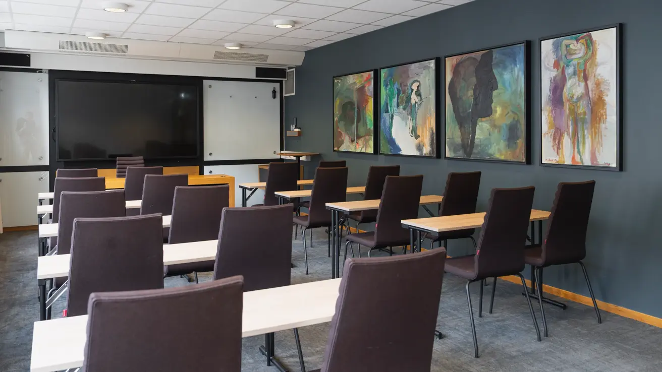 Møterom med klasseroms oppsett, svarte stoler, lyse trebord, mørke grønne vegger og TV skjerm foran.