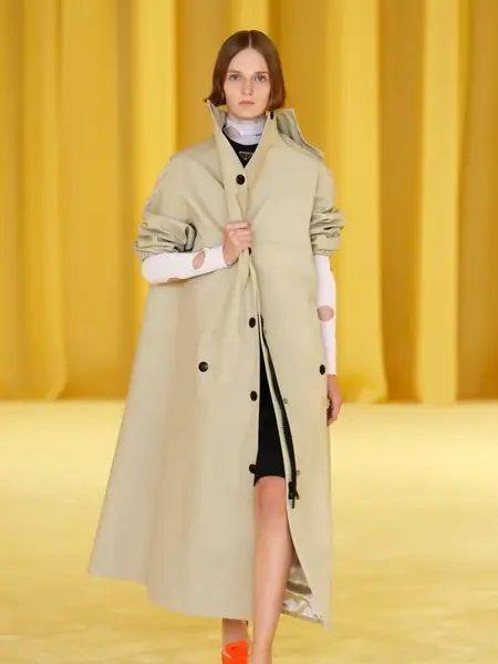 Dame som går på catwalken med veldig gul bakgrunn. Har på seg en beige stor trench coat