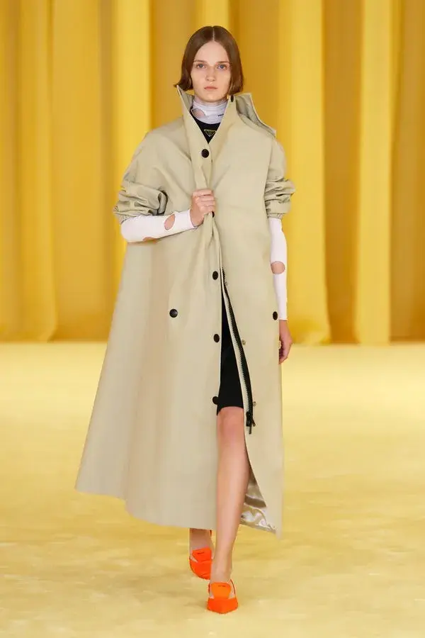 Dame som går på catwalken med veldig gul bakgrunn. Har på seg en beige stor trench coat