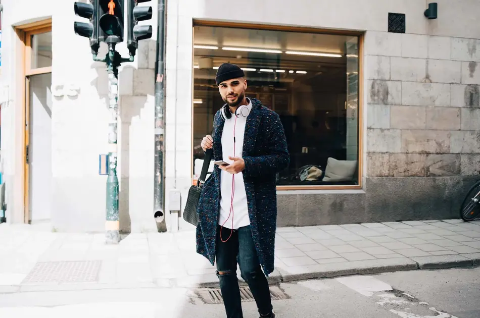 Mann med lang strikkejakke, lys skjorte, hullete skinny jeans, lue og veske med mobil i hånda, stående i urbant landskap