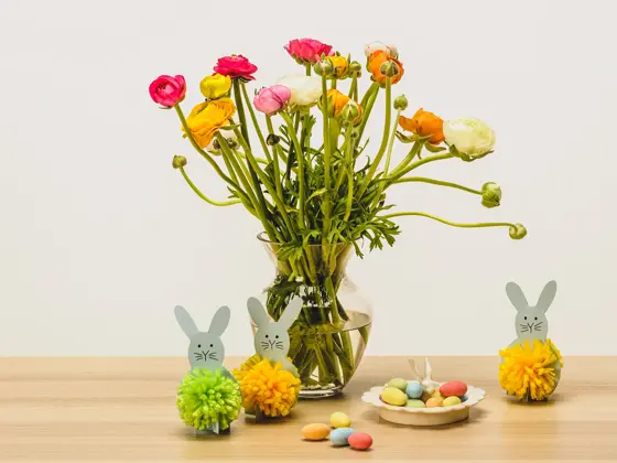 To grå kaniner med dusk laget av grønt og gult garn ved siden av en stor vase med blomster og sjokoladeegg på ett bord