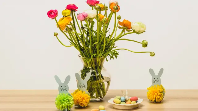 To grå kaniner med dusk laget av grønt og gult garn ved siden av en stor vase med blomster og sjokoladeegg på ett bord