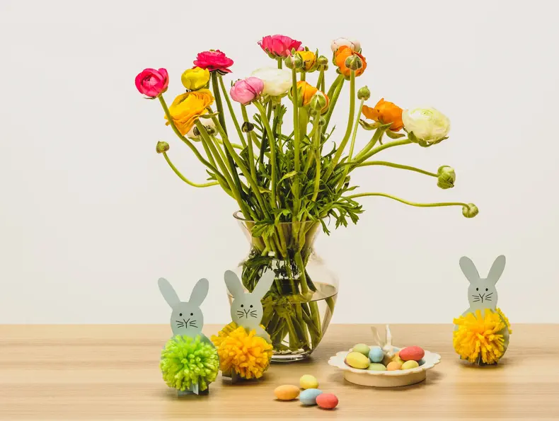 To grå kaniner med dusk laget av grønt og gult garn ved siden av en stor vase med blomster og sjokoladeegg på ett bord