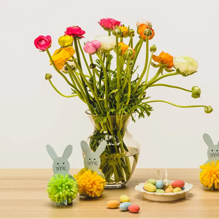 To grå kaniner med dusk laget av grønt og gult garn ved siden av en stor vase med blomster og sjokoladeegg på ett bord