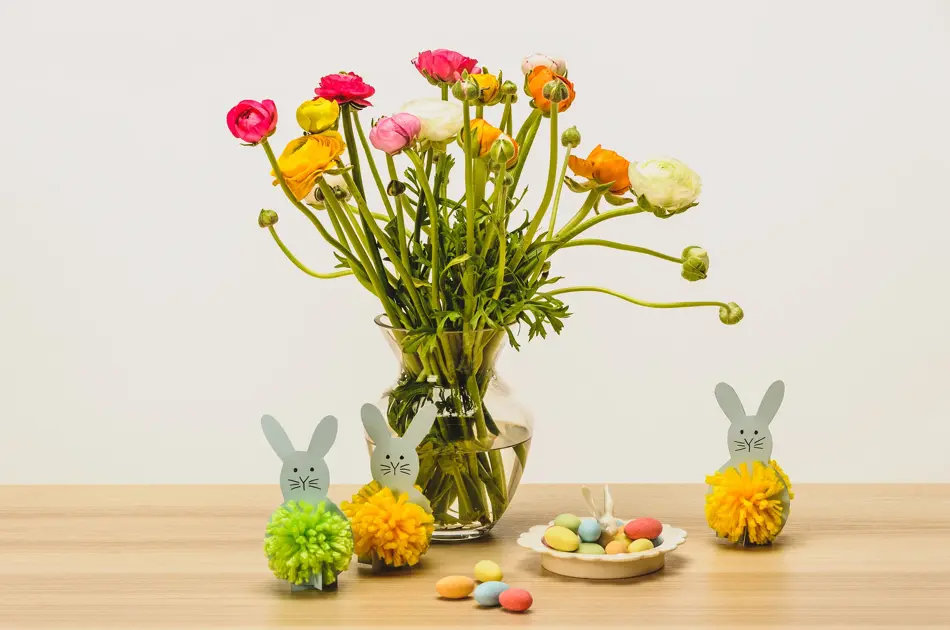 To grå kaniner med dusk laget av grønt og gult garn ved siden av en stor vase med blomster og sjokoladeegg på ett bord