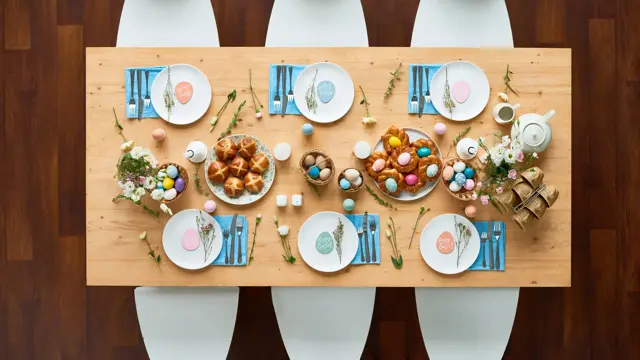 Directly above shot of beautiful Easter table setting: elegant white plates with small egg-shaped greeting cards standing on blue napkins, homemade buns spreading appetizing smell, colorful eggs lying in baskets