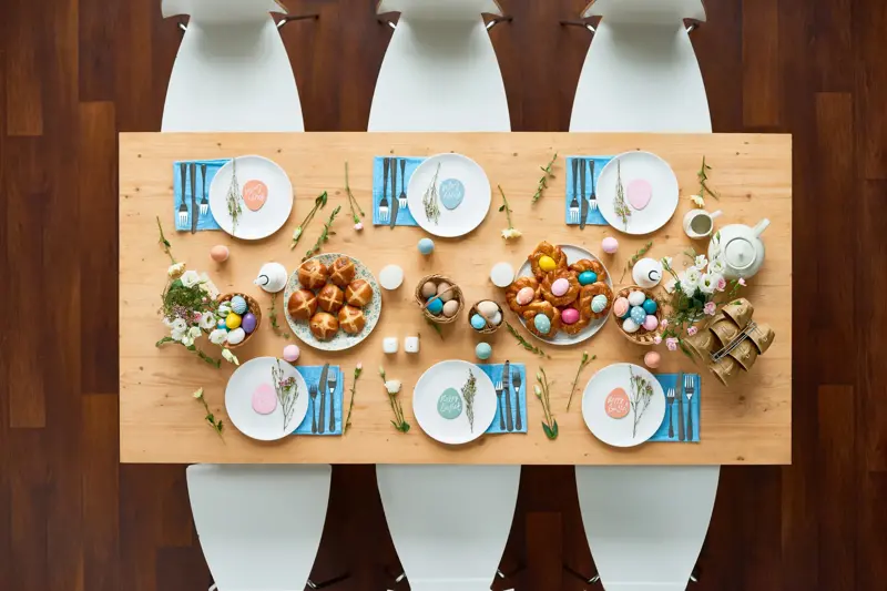 Directly above shot of beautiful Easter table setting: elegant white plates with small egg-shaped greeting cards standing on blue napkins, homemade buns spreading appetizing smell, colorful eggs lying in baskets
