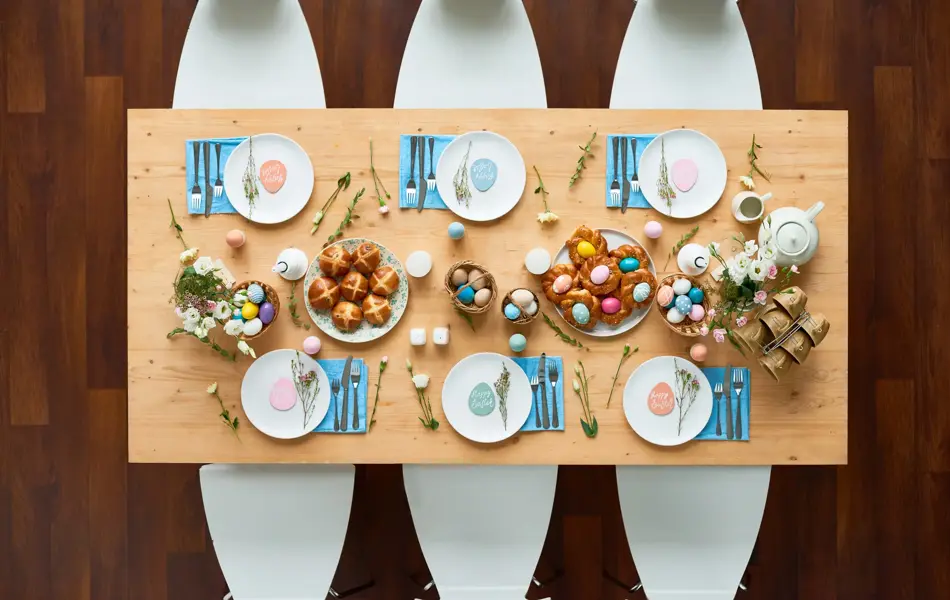 Directly above shot of beautiful Easter table setting: elegant white plates with small egg-shaped greeting cards standing on blue napkins, homemade buns spreading appetizing smell, colorful eggs lying in baskets