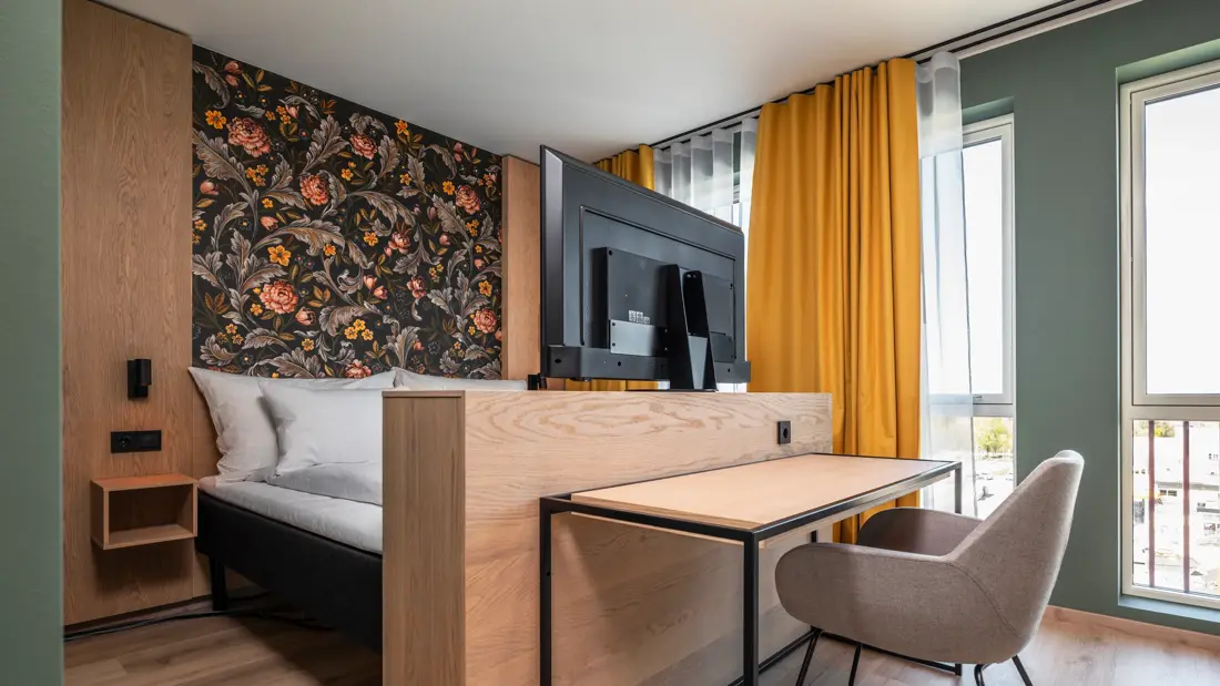 Business rom med dobbeltseng, skrivebord med tilhørende stol, flatskjerm TV, store vinduer med gule gardiner og mønstrete tapet bak sengen.