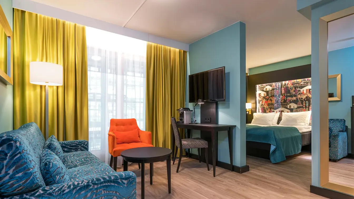 Blå sofa, orange lenestol, skrivebord, smart TV, speil og dobelseng på superiorrom på Thon Hotel Linne
