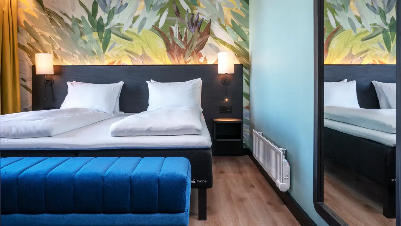 Dobbeltseng, tapetvegg med mønster, blå puff, speil, gule gardiner