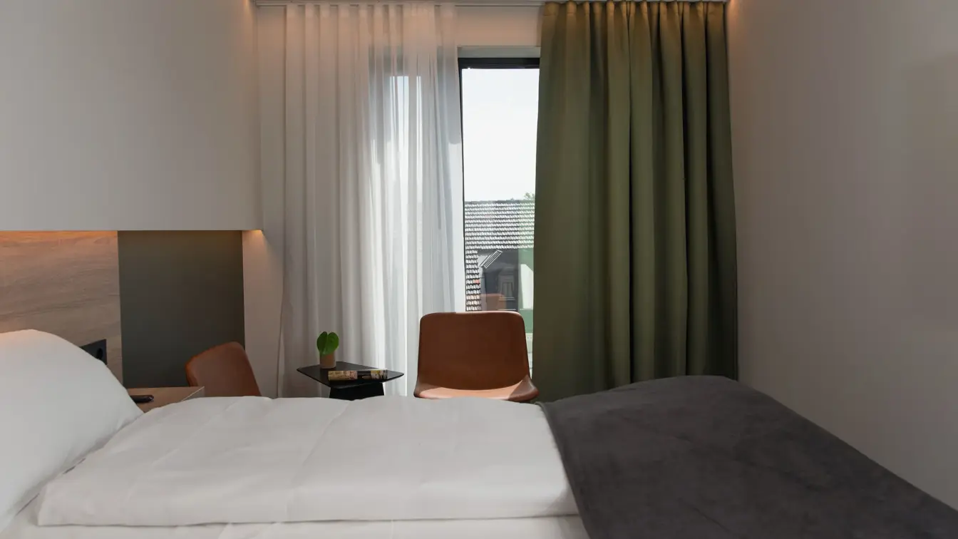 Dobbeltseng med hvite laken og mørkegrå sengeteppe. Stort vindu med grønn gardin. I standard dobbelt rom på Thon Hotel Norge i Kristiansand.