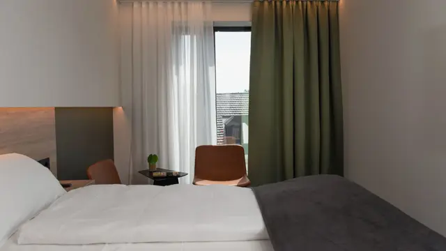 Dobbeltseng med hvite laken og mørkegrå sengeteppe. Stort vindu med grønn gardin. I standard dobbelt rom på Thon Hotel Norge i Kristiansand.