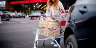 Bildet er tatt utendørs ved kjøpesenteret Charlottenbergs Shoppingcenter og viser en dame i grønn strikkejakke med en handlekurv full av ulike bæreposer.