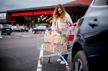 Bildet er tatt utendørs ved kjøpesenteret Charlottenbergs Shoppingcenter og viser en dame i grønn strikkejakke med en handlekurv full av ulike bæreposer.