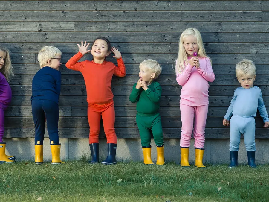 Seks barn står på en gressplen mot en husvegg kledd i ullundertøy og gummistøvler.