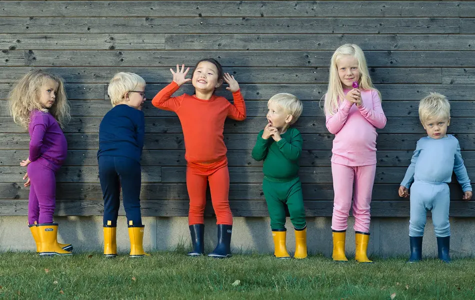 Seks barn står på en gressplen mot en husvegg kledd i ullundertøy og gummistøvler.