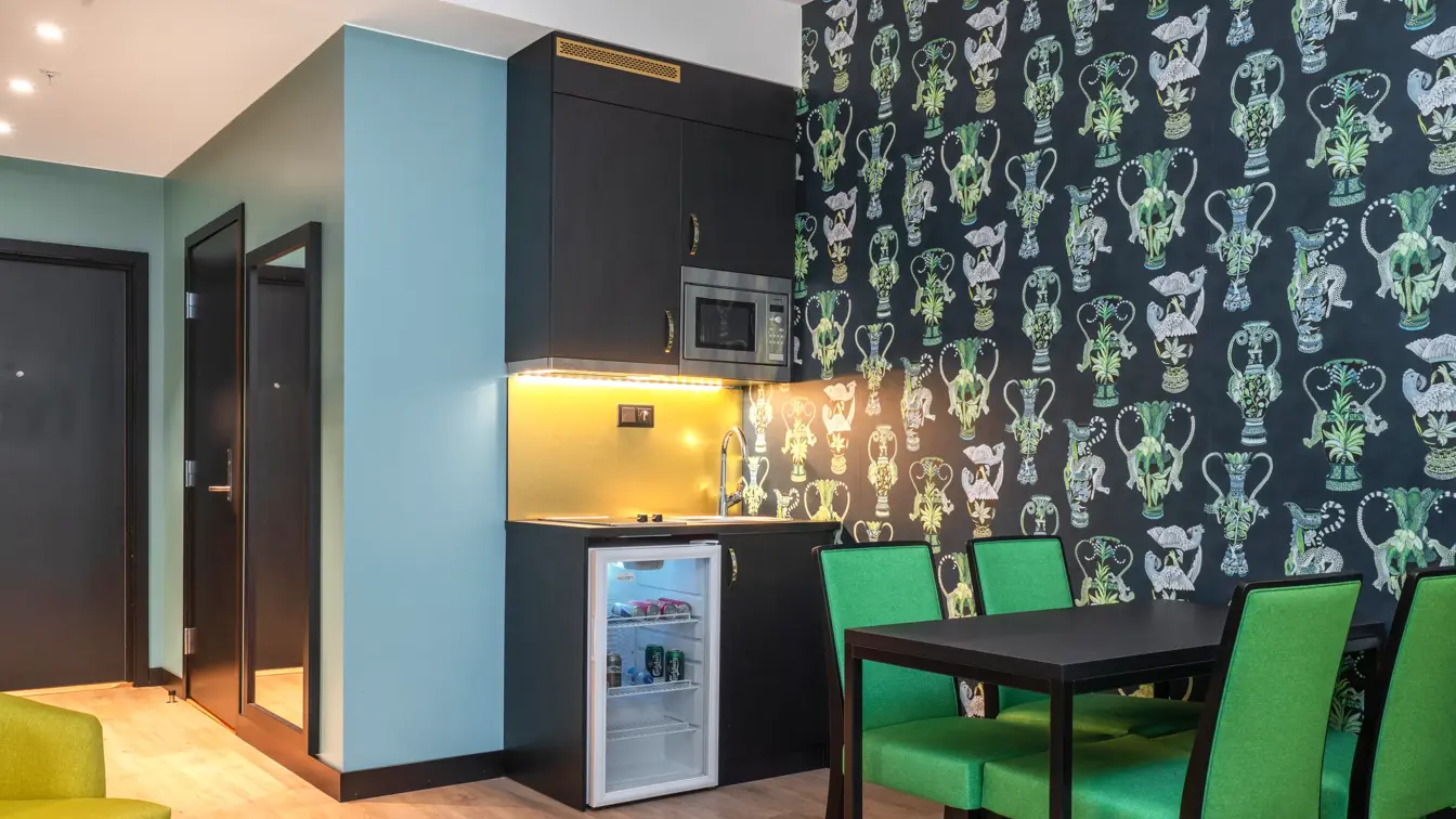 Mørkt spisebord med fire grønne stoler, møstrete tapet med grønne detaljer. Kjøkkenkrokk med spis, vask, større minibar og mikrobølgeovn i junior suite på Thon Hotel Linne.