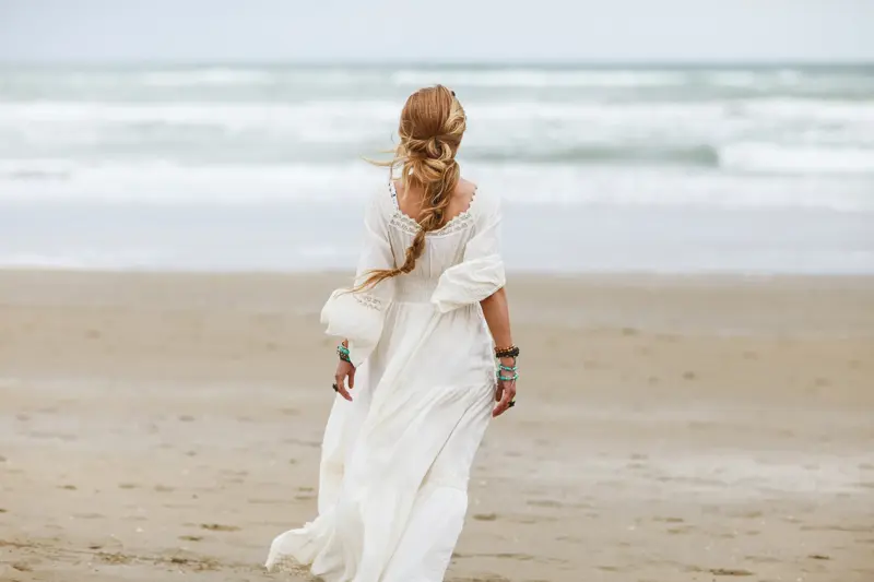Dame med hvit kjole med blonder og volager sett bakfra går mot vannet på strand 