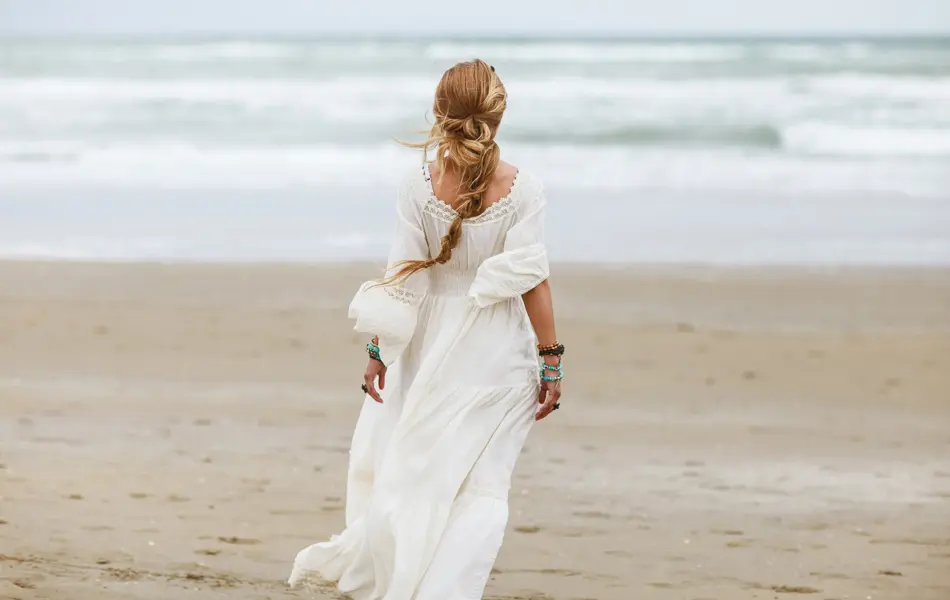 Dame med hvit kjole med blonder og volager sett bakfra går mot vannet på strand 