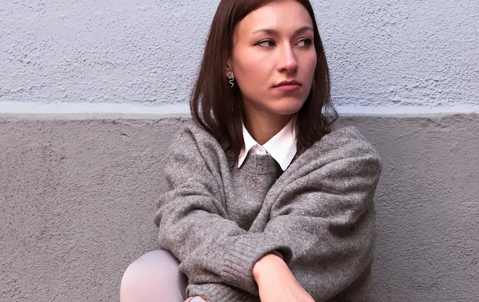 Dame sitter på huk foran en vegg, med grå ullgenser over en hvit skjorte, og sorte boots med metalldetaljer.