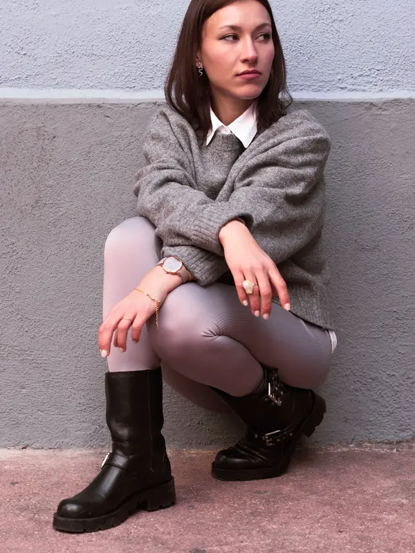Dame sitter på huk foran en vegg, med grå ullgenser over en hvit skjorte, og sorte boots med metalldetaljer.