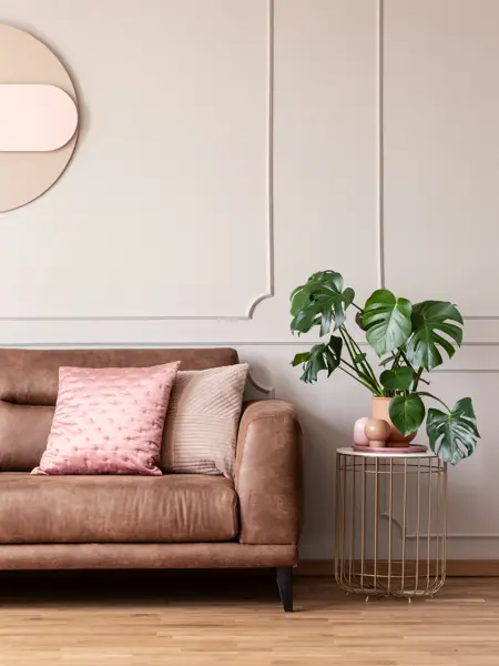 Real photo of end table with decor and fresh plant placed next to leather sofa with pink cushions in light grey living room interior with wainscoting on wall