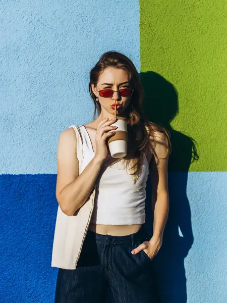 Halvportrett av dame med hvit tanktop, løs, blå bukse og røde solbriller som drikker av kopp med sugerør står inntil fargerik murvegg.  