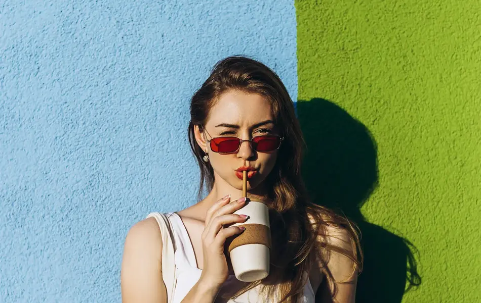 Halvportrett av dame med hvit tanktop, løs, blå bukse og røde solbriller som drikker av kopp med sugerør står inntil fargerik murvegg.  